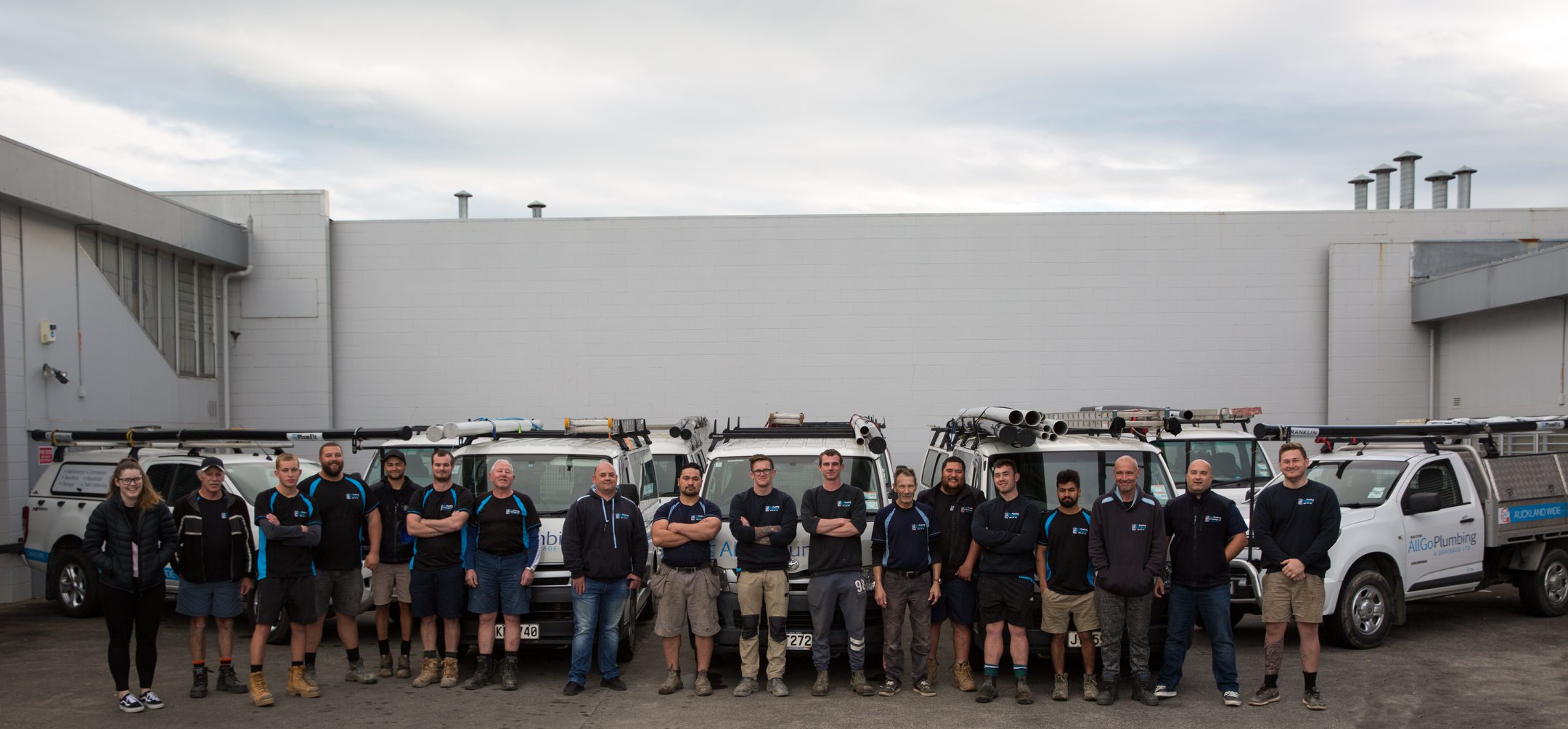 AllGo Plumbing Team with Fleet – Auckland’s Trusted Local Plumbers A group of 20+ people from AllGo Plumbing standing in a row in front of a fleet of branded white plumbing service vehicles. The team includes both men and women wearing company uniforms. The vehicles are equipped with ladders and plumbing equipment, and the setting appears to be the company’s garage or parking area in Auckland.